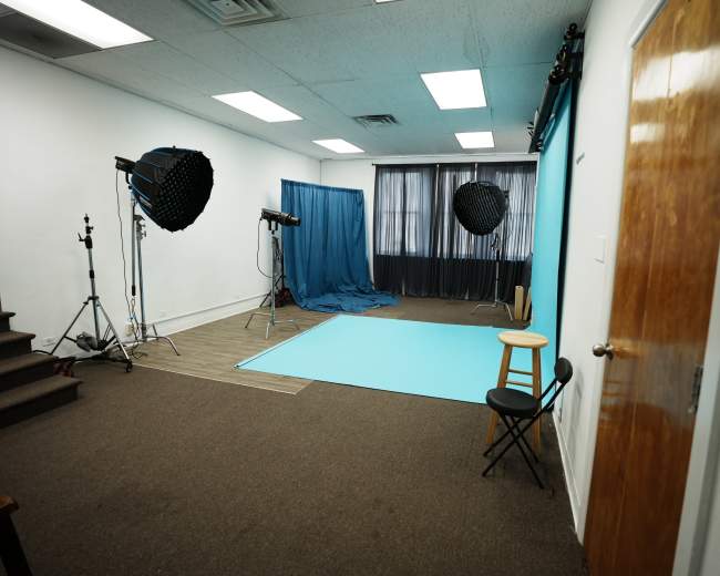 The image shows a photography studio featuring a blue backdrop, two light setups, a blue curtain, and a stool positioned in the corner.