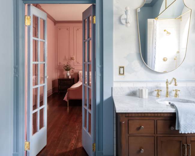 A bathroom features a double vanity with a large mirror, leading to a room adorned in soft pink hues.