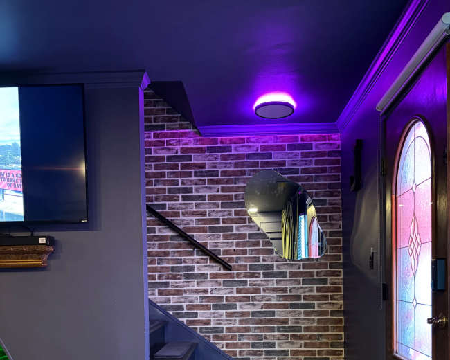 The image shows a modern entryway featuring a staircase with a brick accent wall, a round mirror, a television, and colorful lighting.