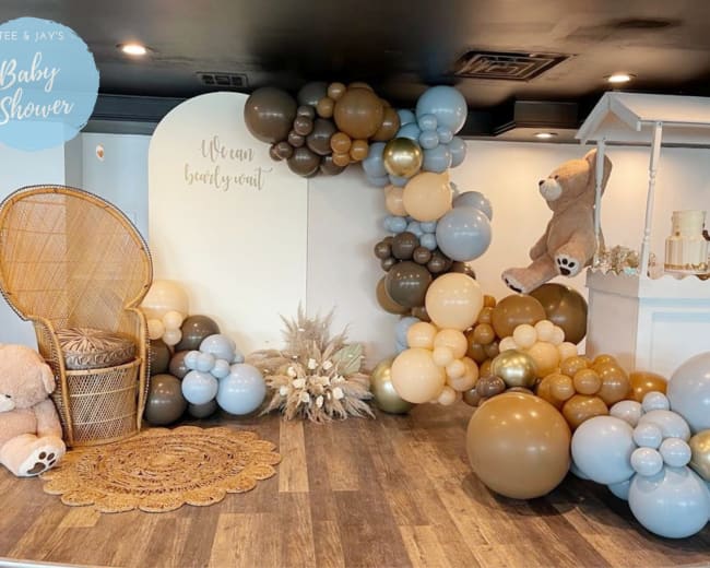 A baby shower setup with a decorative arrangement of balloons in blue, beige, and brown, accompanied by a plush teddy bear and a round, woven mat on a wooden floor.