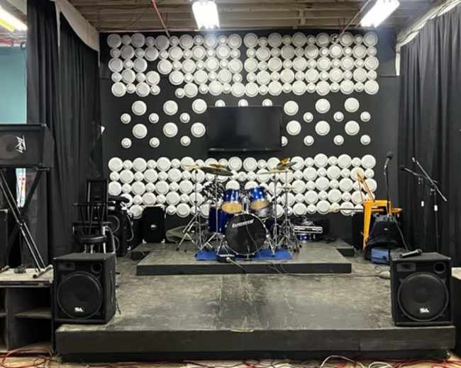 The image shows a stage setup with a drum kit, amplifiers, and instruments against a backdrop of white circular decorations.