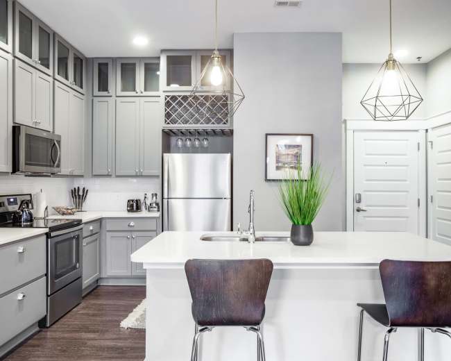 The image shows a modern kitchen with gray cabinetry, stainless steel appliances, a white countertop, and two bar stools.