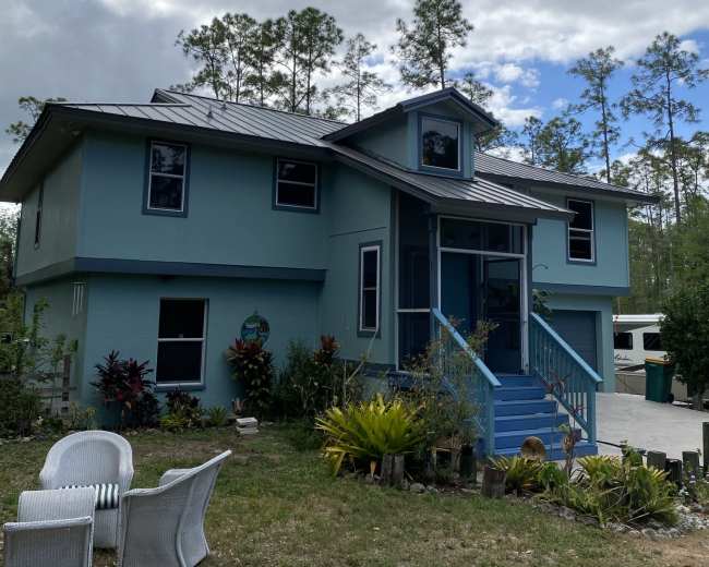 A two-story blue house with a metal roof and a front porch surrounded by greenery and decorative plants.