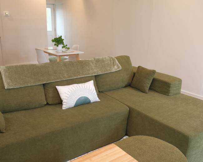 A green sectional sofa with a decorative pillow is positioned in a bright, minimalist living space that features a dining table in the background.