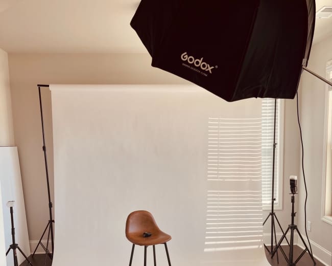 A photography studio setup featuring a brown stool in front of a white backdrop, illuminated by softbox lights on either side.