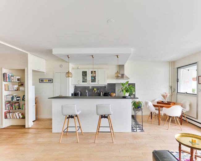 The image shows a modern open-concept kitchen and living area with bar stools, a dining table, and large windows.