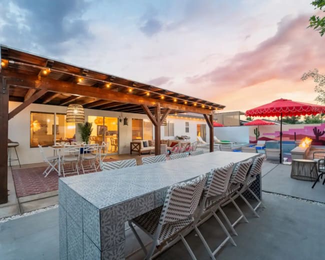 The image shows an outdoor patio with a long dining table, surrounded by chairs, under a wooden pergola adorned with string lights, featuring a swimming pool and colorful umbrella in the background.