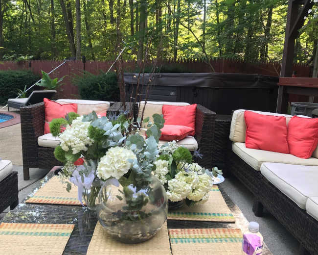 A patio area features wicker furniture with bright cushions, a glass table adorned with a large floral arrangement, and a hot tub in the background surrounded by greenery.