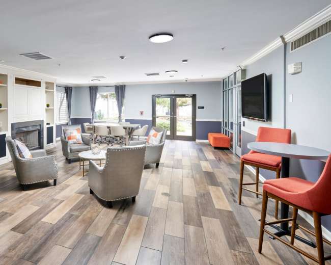 The image shows a modern lounge area with a seating arrangement of chairs and a table, surrounded by large windows and light-colored walls.