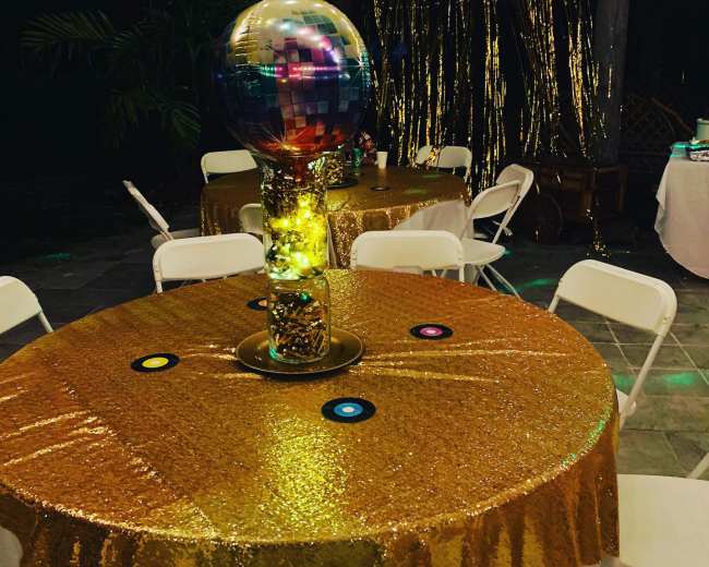 The image shows a decorated table with a gold sequin tablecloth, featuring a disco ball centerpiece and colored vinyl record accents.