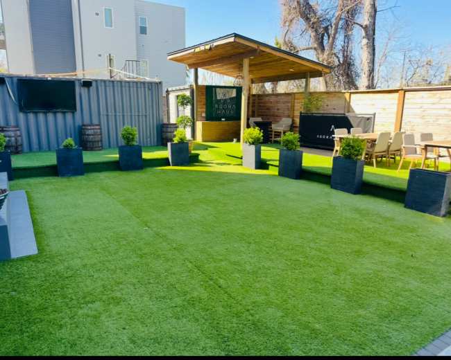 The image shows a landscaped outdoor space with artificial grass, potted plants, and a wooden gazebo featuring a seating area.
