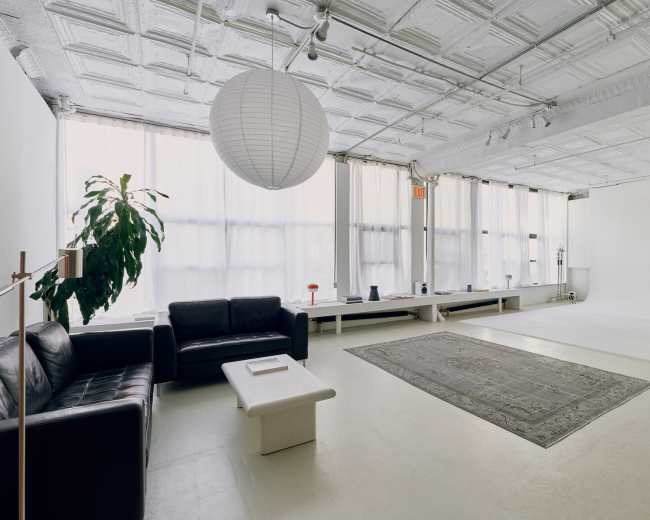 A minimalist studio space features a black leather sofa, a light-colored coffee table, and a large area rug on a polished floor, with ample natural light coming through sheer curtains.
