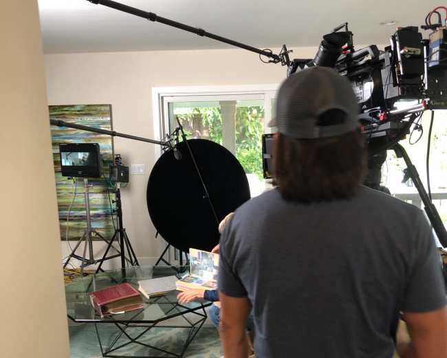 A crew member stands in a living room set up for filming, surrounded by cameras, lights, and a reflector.