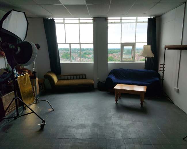 The image shows a sparsely furnished studio space with large windows, a couch covered with a blue blanket, a wooden table, and lighting equipment set up.