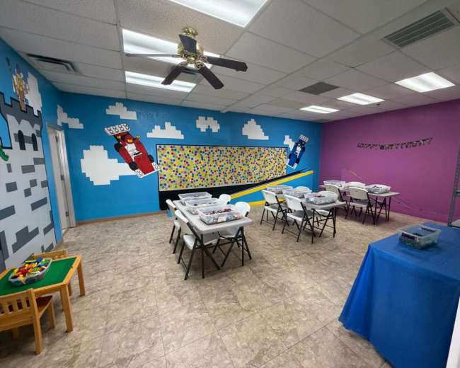 The image shows a brightly colored classroom with a large mural on one wall, several folding tables with chairs set up, and shelves along one side.