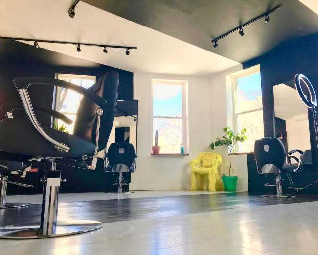 The image shows a modern hair salon with several black styling chairs, large windows, and a yellow armchair in the corner.