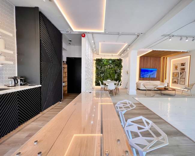 The image shows a modern interior space featuring a kitchen area with a long wooden table and white chairs, leading to a lounge area with a television and greenery.