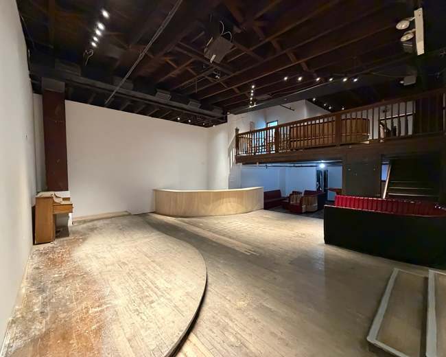 The image shows an empty auditorium with a wooden stage, a raised balcony, and sparse seating on the lower level.