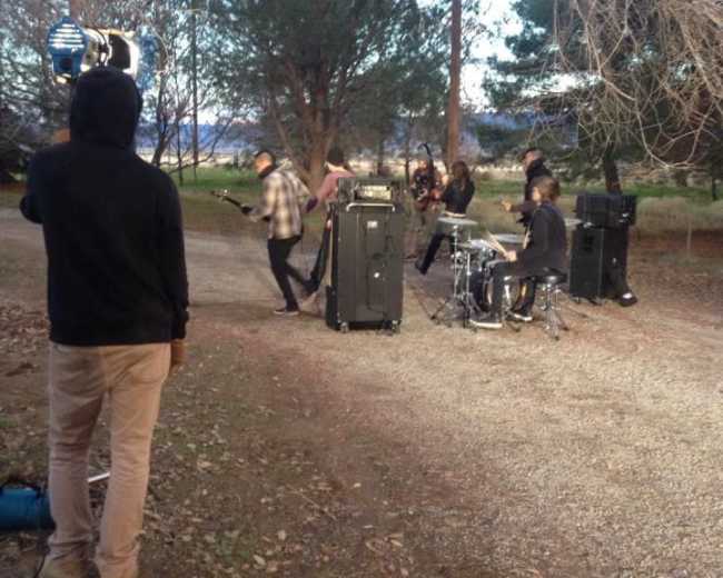 A band performs in a wooded area, with a drummer and several guitarists set up near musical equipment on a gravel path.
