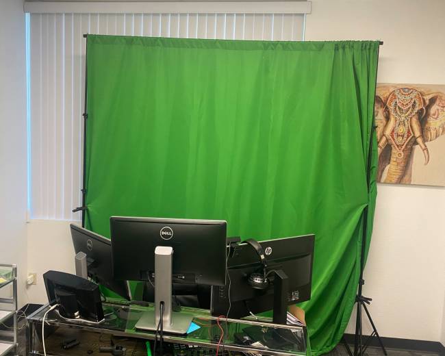 A workspace featuring a green screen setup behind dual computer monitors, with a decorative elephant painting on the wall.