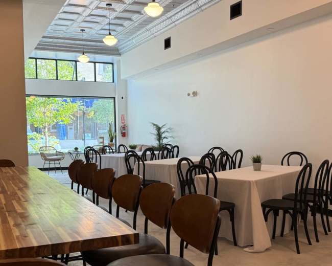 The image shows an empty dining area with wooden and metal chairs, tables covered with white linens, and a large window letting in natural light.
