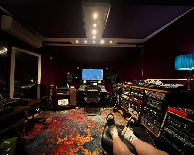 A music recording studio features an array of audio equipment, a central mixing console, and colorful carpet, with a person’s legs visible in the foreground.