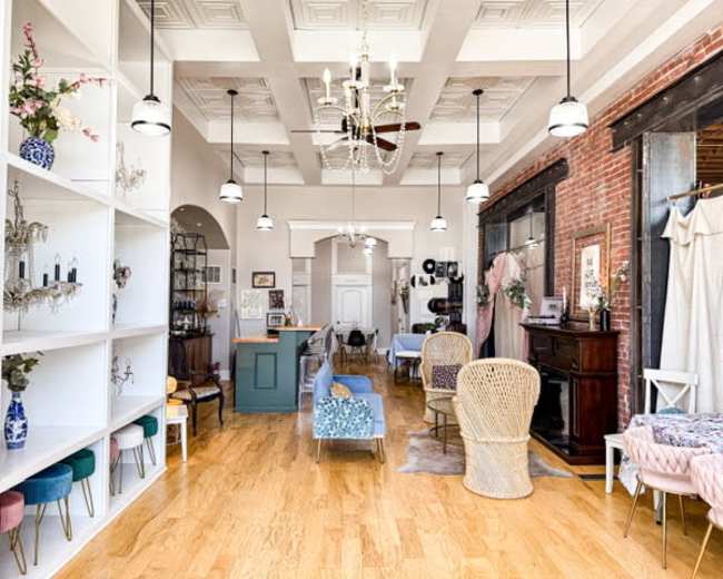 The image shows a stylish interior of a boutique or event space with wooden flooring, decorative furnishings, and high ceilings featuring hanging light fixtures.