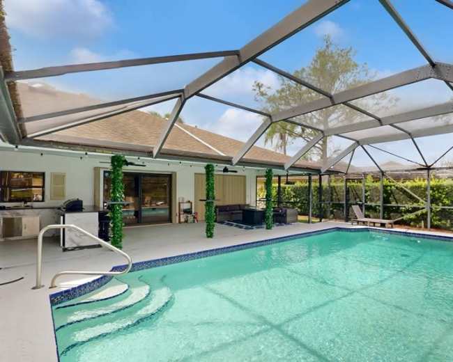 The image shows a swimming pool enclosed by a netted structure, with a clear blue sky and surrounding greenery visible in the background.