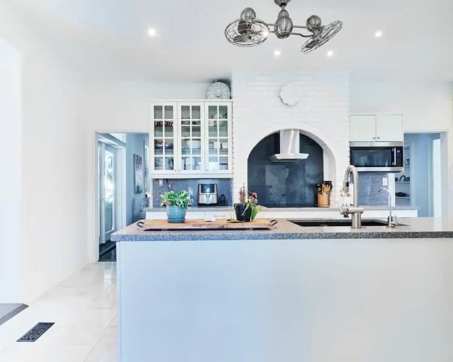 The image shows a modern kitchen with a central island, white cabinetry, and a slate grey countertop featuring a built-in stovetop and hood.
