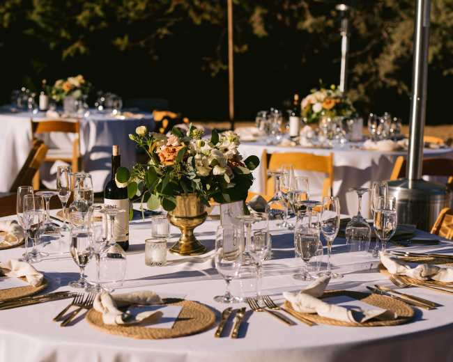 A beautifully arranged outdoor dining setup features round tables with white tablecloths, elegant glassware, and floral centerpieces.
