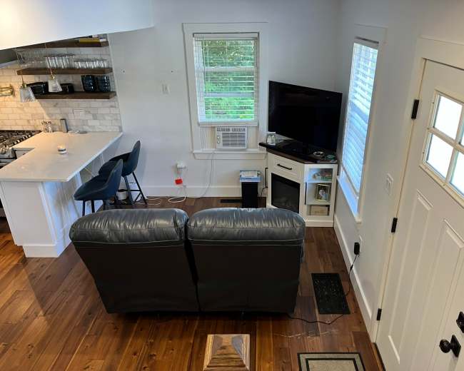 The image shows a cozy living space with a gray leather couch facing a television, a side table, and a door leading outside.