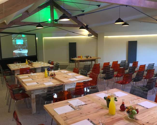 A spacious meeting room is set up with several wooden tables arranged for dining, surrounded by colorful chairs and featuring a projection screen at one end.
