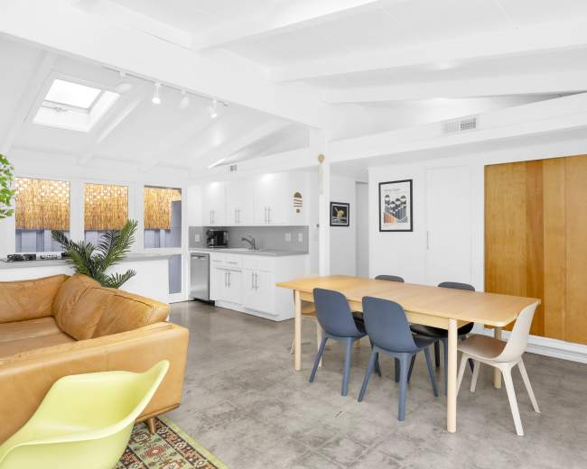 The image shows a modern living space with a kitchen area, dining table, and a couch, featuring bright light from overhead windows and large wooden sliding doors.