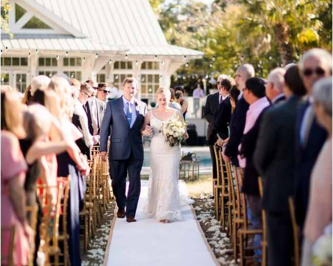 A bride and groom walk down a floral-decorated aisle surrounded by seated guests at an outdoor wedding ceremony.
