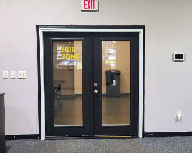 The image shows a set of double glass doors labeled "THE HUB ZONE," with an exit sign above and a visible room behind them.