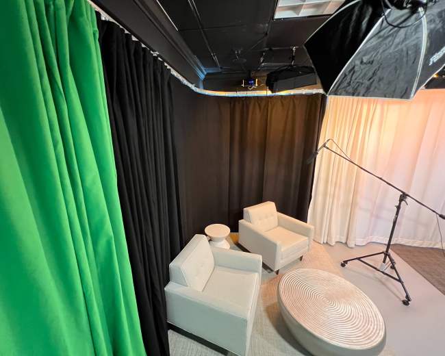 Two light gray chairs positioned near a round white table against a backdrop of green, black, and white curtains in a studio setting.