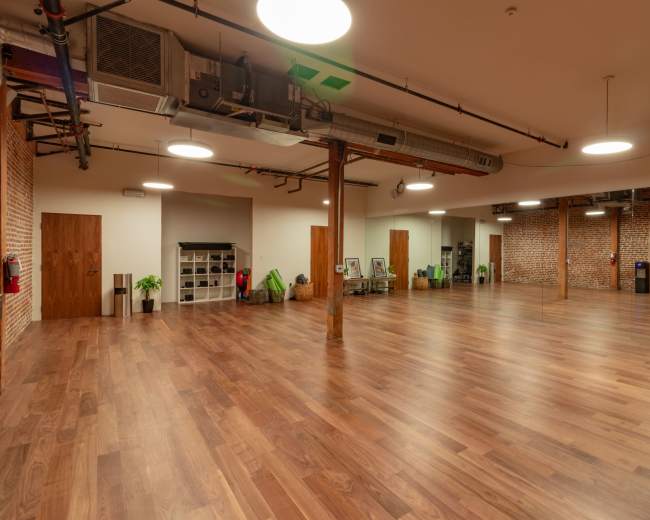 The image shows a spacious, empty interior room with hardwood flooring, exposed brick walls, and bright overhead lighting.