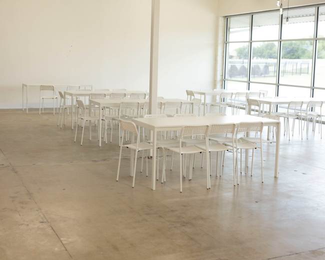 The image shows a spacious, empty dining area with several white tables and chairs arranged neatly, and large windows allowing natural light to enter.