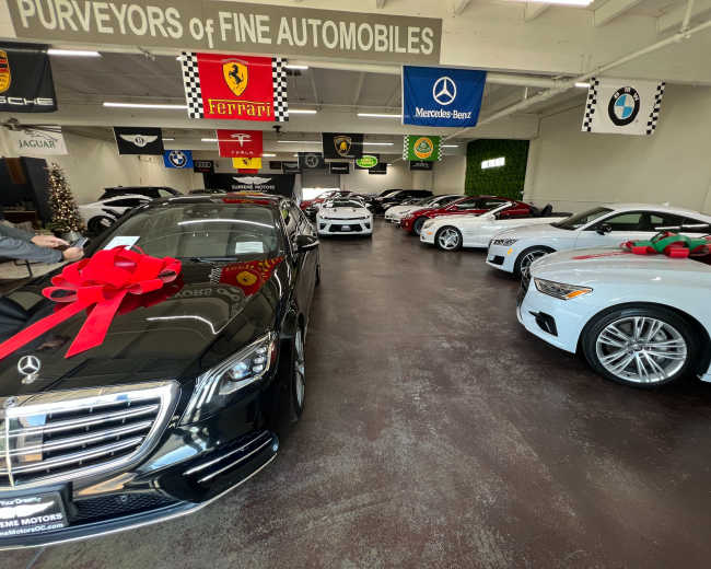 The image shows a car showroom featuring a lineup of various luxury vehicles, including brands like Mercedes-Benz, BMW, and Audi, adorned with festive decorations and large bows.