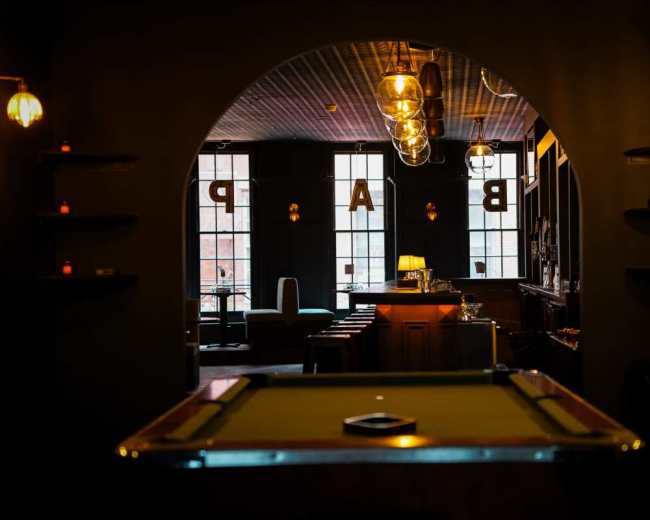 The image shows an interior scene of a bar, featuring a pool table in the foreground and dimly lit seating areas with large windows in the background.