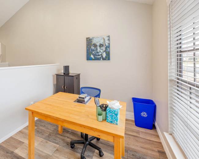 The image shows a small office space featuring a wooden desk, a chair, a black cabinet, a trash bin, and a colorful painting on the wall.