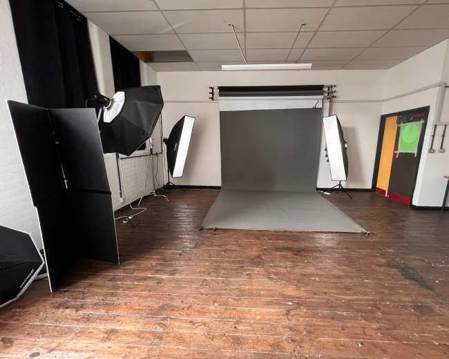 The image shows a photography studio with a gray backdrop, lighting equipment positioned around, and wooden flooring.