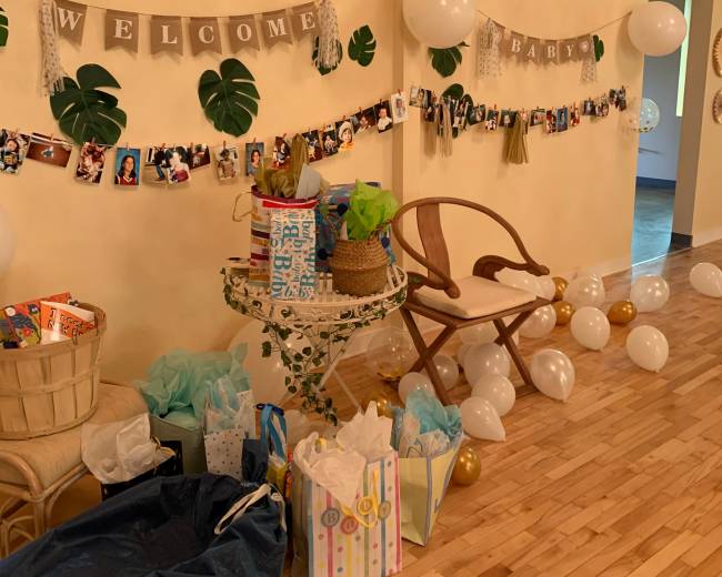 A decorated wall with a "Welcome Baby" banner, featuring photos and balloons, is surrounded by a table stacked with gifts and bags.
