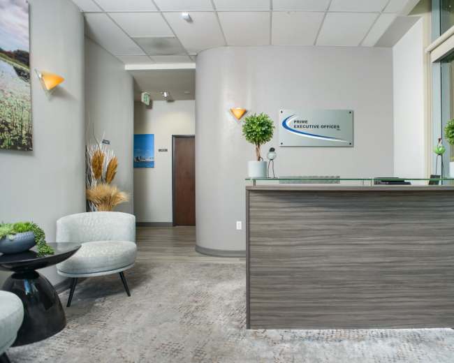 The image shows a modern office reception area with a wooden front desk, two light-colored chairs, and decorative plants, along with a sign for "Prime Executive Offices" on the wall.
