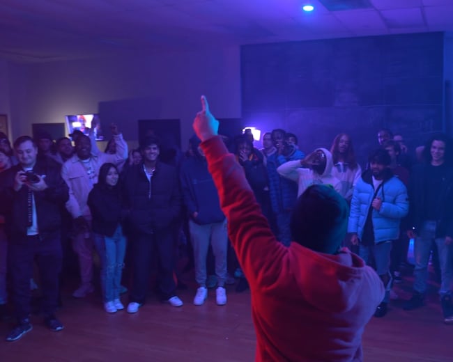 A crowd of people gathers in a dimly lit room as a performer gestures with one hand raised while standing in front of them.