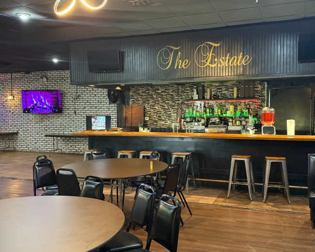 The image shows a bar area with a dark wooden counter, several high stools, and a prominently displayed sign reading "The Estate," along with illuminated shelves stocked with various bottles.