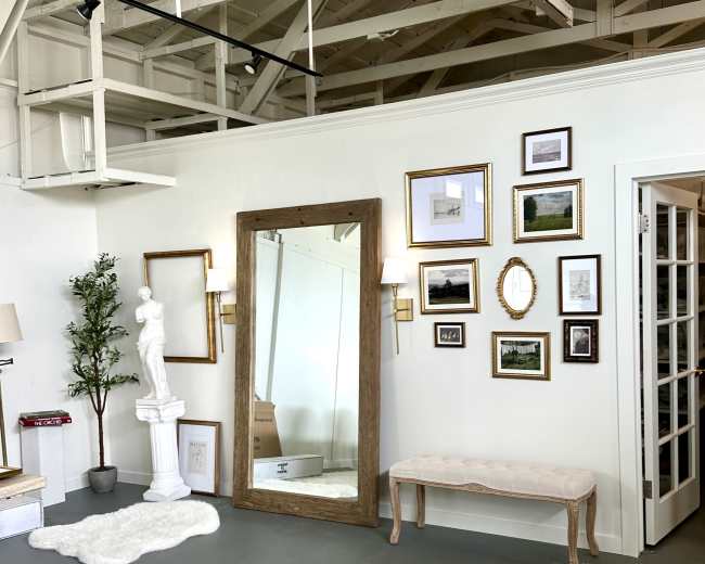 The image shows a spacious room with a large mirror, various framed pictures on the wall, a statue, and a bench in front of a glass door.
