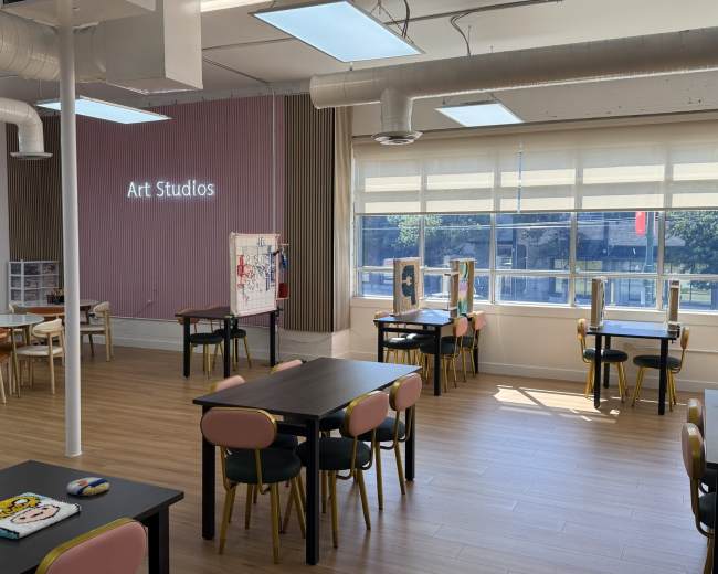 The image shows a well-lit art studio with several tables and chairs arranged throughout the space, featuring large windows that provide natural light.