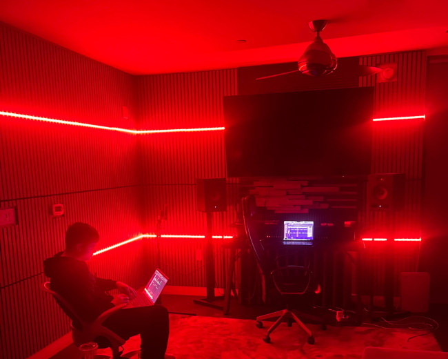 A person sits in a chair with a laptop in a room illuminated by red LED lights, surrounded by audio equipment.