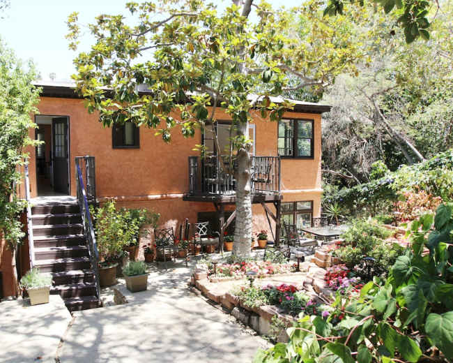A two-story house with orange walls and black railings is surrounded by a garden featuring potted plants and flowers.